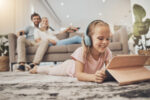 A girl lays on a living room rug, listening to an audiobook with headphones. Her parents are sitting behind her on the couch, looking on.