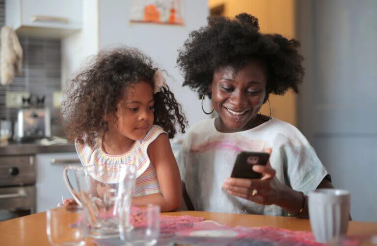 A young girl watches as her mom smiles toward her cell phone.