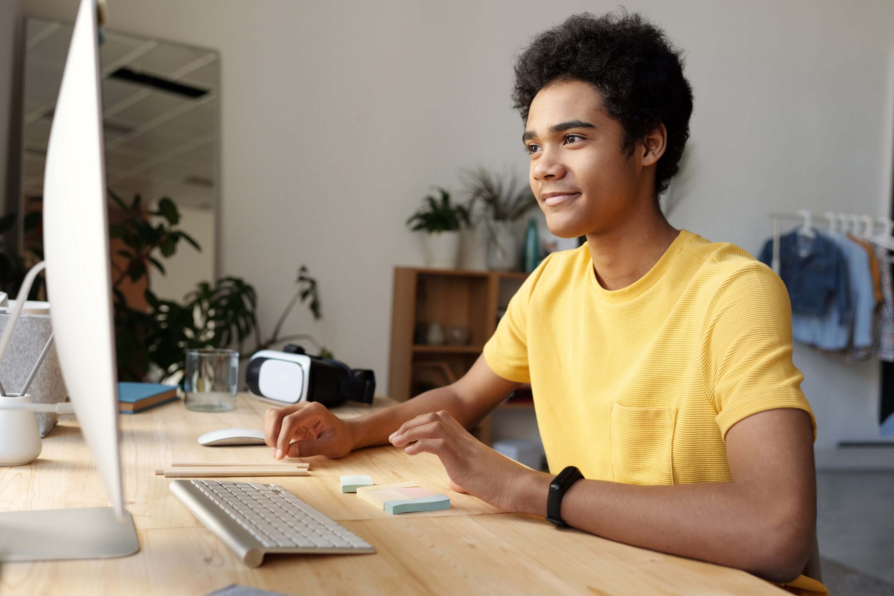 A teen male in a yellow shirt sits in front of his computer at home.