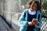 Young girl leans against a fence at school while looking at her cell phone.