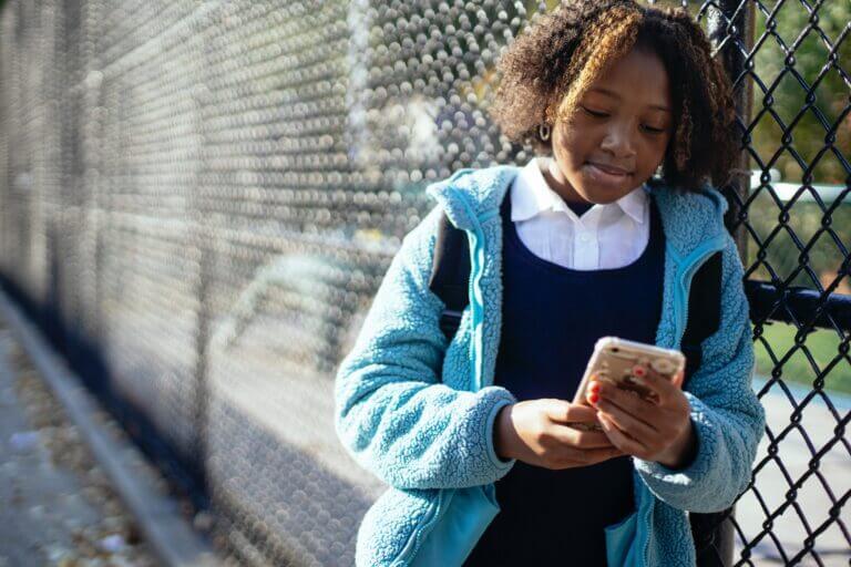 Young girl leans against a fence at school while looking at her cell phone.