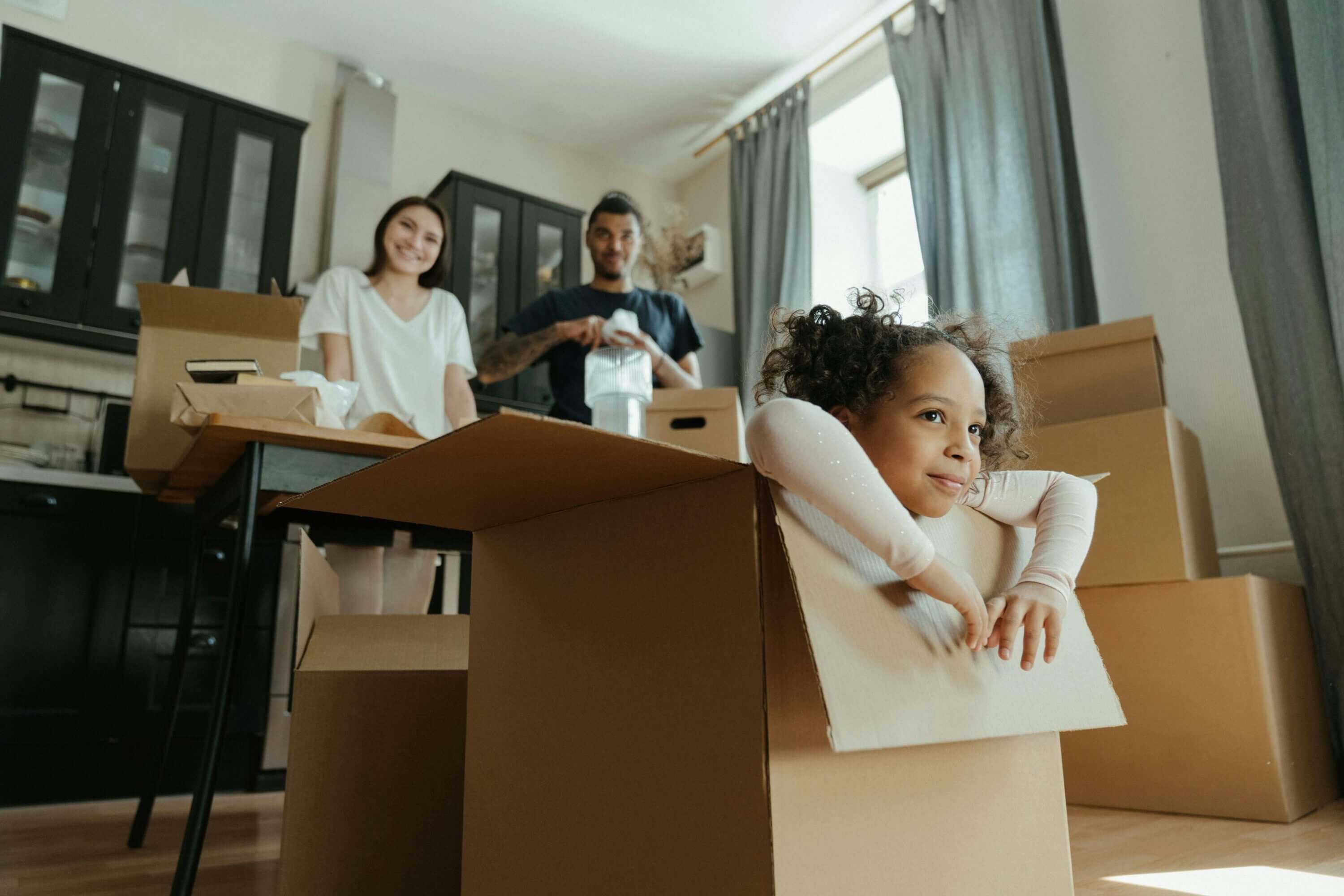 A young girl hangs out of a moving box while her parents unpack at their new house.