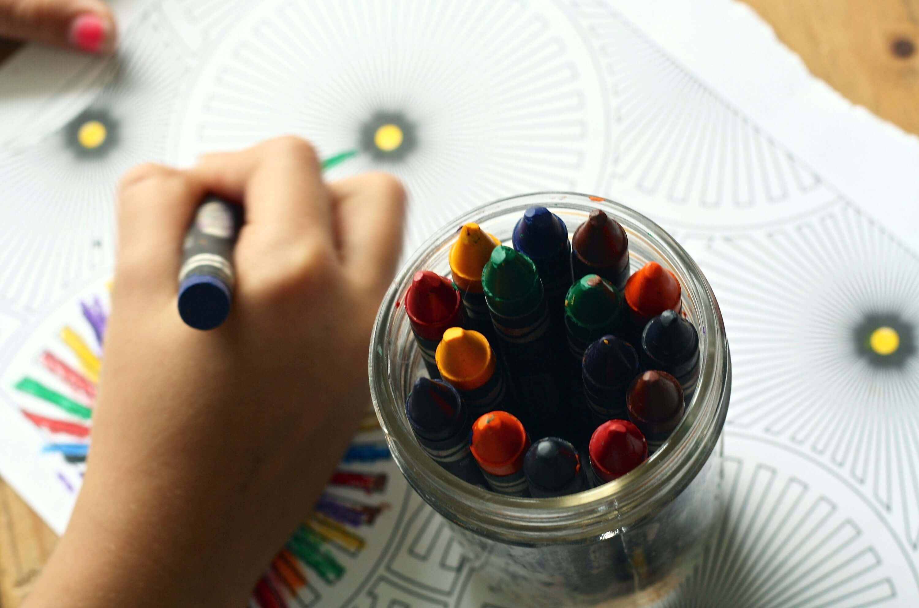 A child colors a printed coloring sheet with a cup of crayons next to their hand.