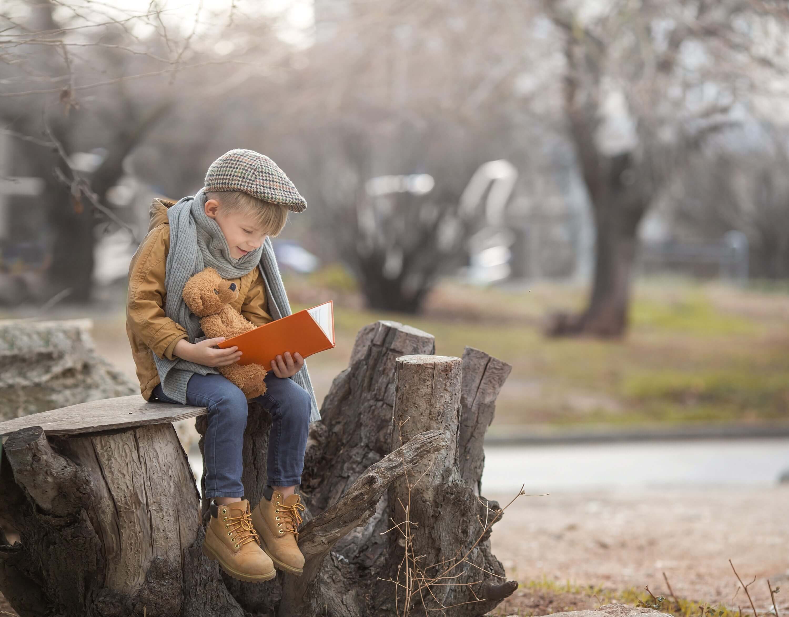 A young boy sits on a tree stump with his stuffed animal, reading a book.