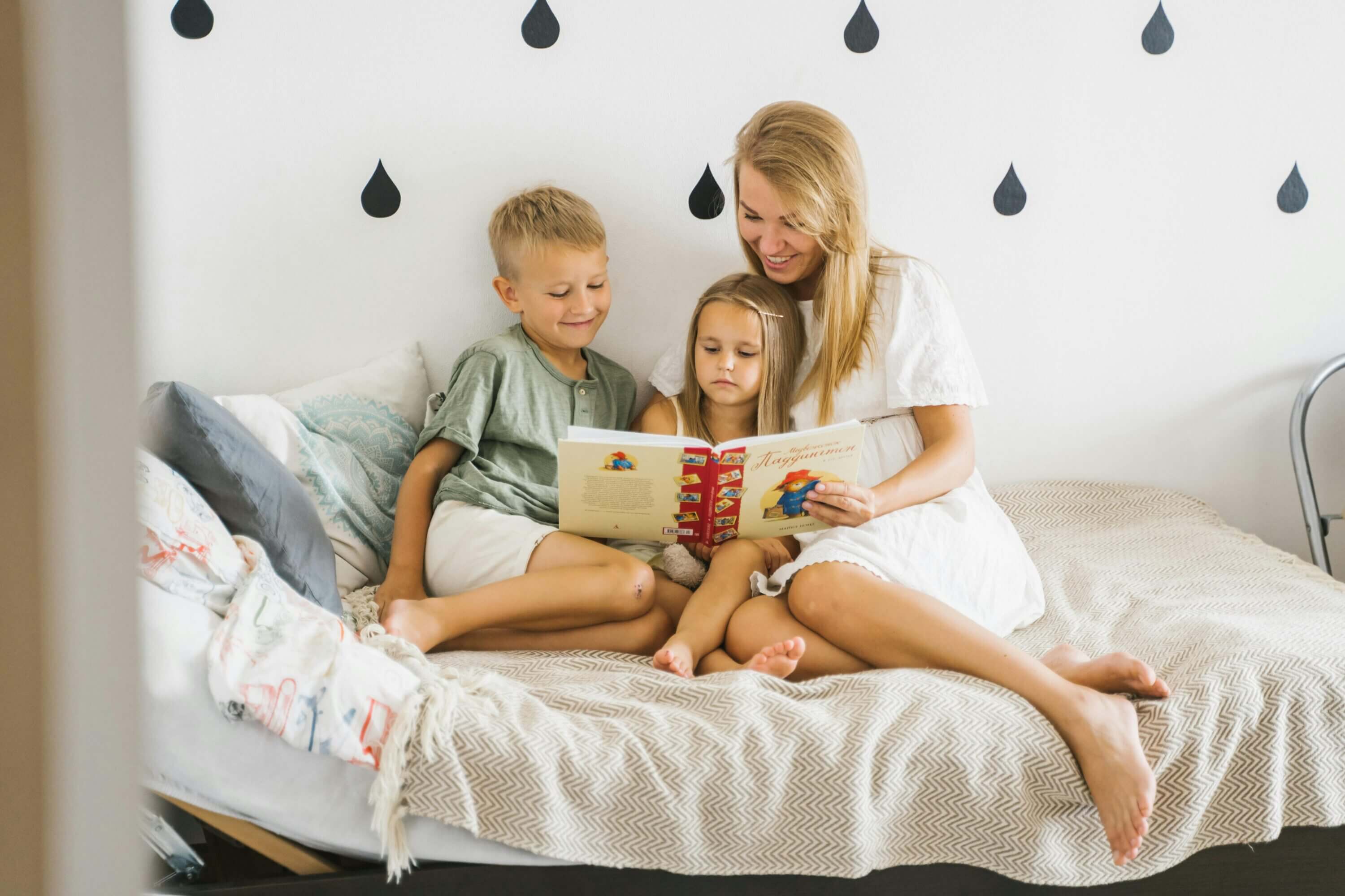 A mom and two kids sit on a bed and read a story together.