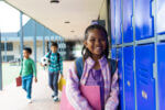 A young girl holds a pink folder and wears a blue backpack while leaning against lockers at her school.