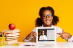 A young girl in classes shows off her school lesson on a tablet. Sitting next to her is a stack of books with an apple on top.